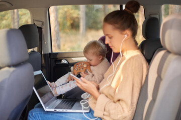 Portrait of Caucasian beautiful woman working on notebook while sitting with her baby daughter in safety chair on backseat of car, female listening music on phone and typing on laptop.