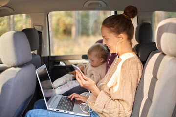Profile portrait of dark haired smiling satisfied woman working on laptop while sitting with her baby daughter in safety chair on backseat of car, young adult girl using cell phone and listening music