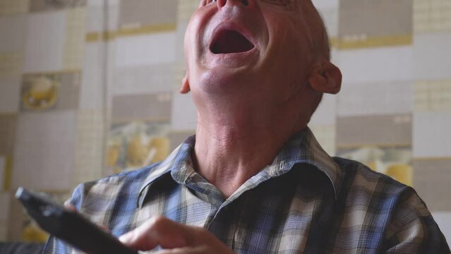 funny pensioner with open mouth and surprise with a remote control in his hands watches tv at home. TV programs for the elderly. senile dementia, pensioner enjoys watching tv