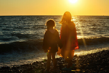 two little girls stand in the evening on the beach with their backs to us in the rays of the sun and watch a beautiful sunset on the seashore, selective focus, silhouettes of people