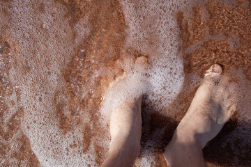 Top view of men's legs in the sea. A man is standing on the sand. An ocean wave washes the shore. Summer holidays.