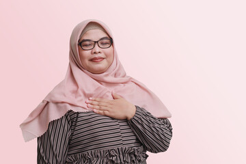 Portrait of cheerful Asian woman with hijab, placing hand on her chest and feeling peaceful. Mental...
