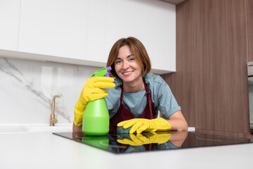 Young attractive and smiling housewife in red apron and yellow rubber gloves cleaning table with rag. Household and cleaning service concept