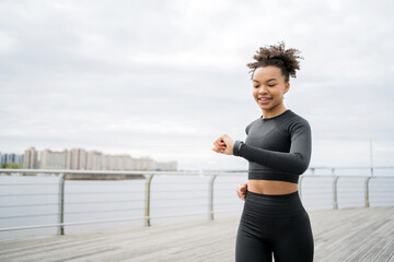 Looks at the fitness watch, a woman runs, smiles, trains fitness in a tight-fitting tracksuit.