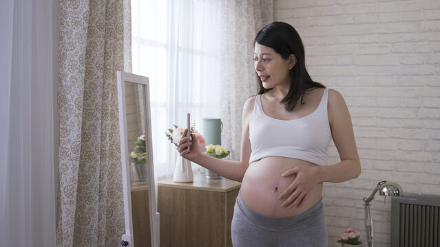 joyful asian lady in the third trimester is holding her belly while taking a mirror selfie with her mobile phone in the bright home interior.