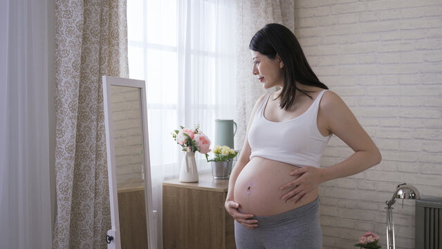 chinese lady in the last trimester standing by a mirror is touching and looking at her belly in different angles with joy at bright home interior.