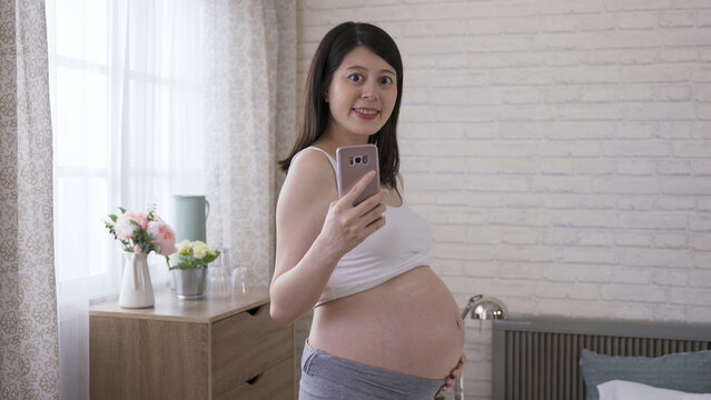 happy pregnant mother facing camera and holding a mobile phone is taking a mirror selfie at home with white brick wall in the background.