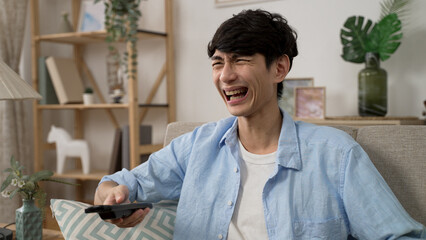 closeup of a cheerful asian male holding remote control is bursting out laughing while watching a comedy on tv in the living room at home