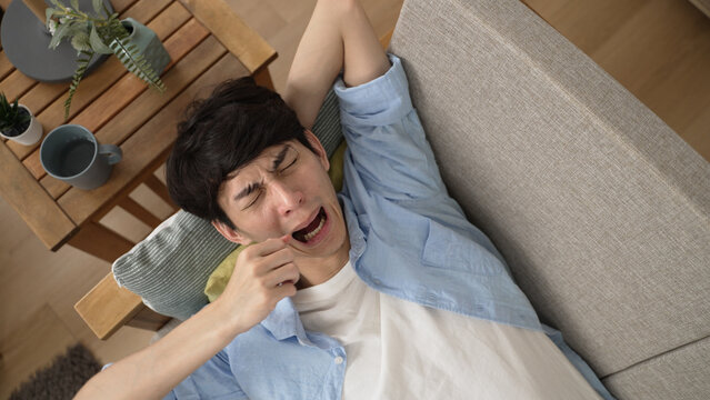 top angle view of a bored Asian japanese man yawning with a stretch and rubbing his face while reclining lazily on the sofa in the living room at home during isolation