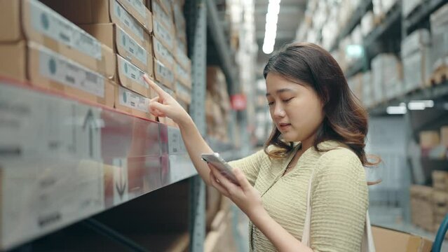 Asian woman using smartphone shopping on shelves at large warehouse retail store. Rack of furniture and home accessories store. Interior of cargo in eCommerce and logistic concept.