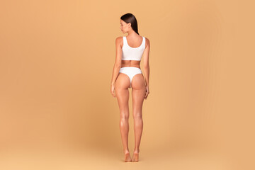 Back view of slim woman with perfect figure posing in white underwear over beige studio background,...