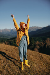 Woman fall smile with teeth full-length hands up happiness walking on the hill and looking at the mountains in a yellow raincoat and jeans happy sunset trip on a hike, freedom lifestyle 