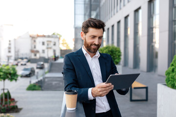 Happy confident young caucasian guy with beard in suit typing on tablet on balcony of modern office...