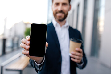 Cheerful confident young caucasian guy with beard in suit shows phone with blank screen, hold cup of coffee