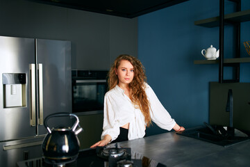 Portrait of blonde caucasian woman standing and smiling in her kitchen leaning on the table. Young...