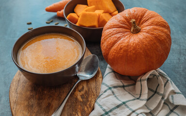 Pumpkin cream soup in a bowl with a spoon on wooden board on the table next to pumpkin whole and cut into pieces.  Healthy, vegetarian food. Diet. Home cooking. Thanksgiving day. Halloween.