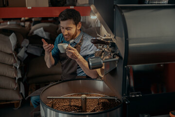 Male barista inhaling aroma of fresh made coffee on the background of roasting coffee machine