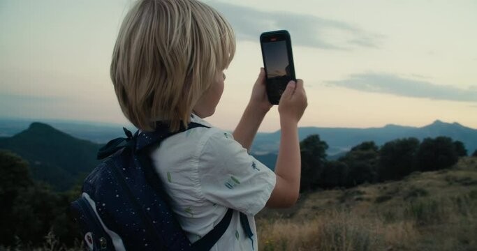 Children on vacation travel with backpack on mountain top at sunset using smartphone. Child boy photographing on smartphone during vacation journey adventure. 