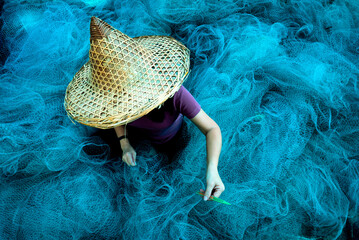 Women fisherman hands sewing blue fishing nets sitting on the ground and surrounded big net