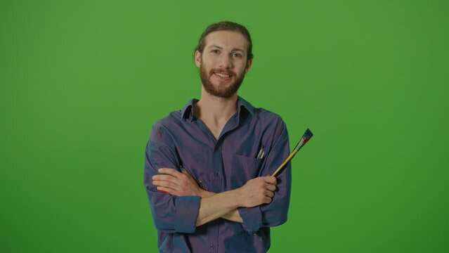 Portrait of Talented Young Bearded Artist, with Dirty Paint, on an Apron, Crosses His Arms, Holding Brushes, Looks at the Camera Smiling on a Green Screen,Chroma Key.The Creativity And Art Concept.