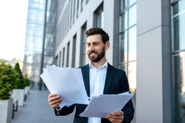 Happy busy handsome young caucasian guy with beard in suit works with documents on street