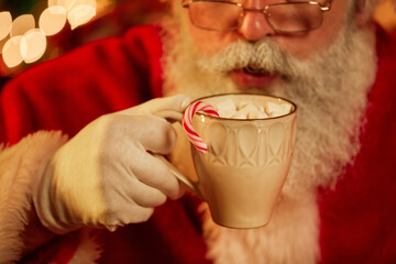 Close up of traditional Santa Claus enjoying cup of hot chocolate with marshmallows and Christmas...