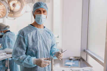 Fototapeta na wymiar Professional surgeon in mask and gloves standing with surgical instruments in operating room