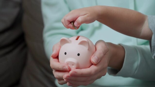 The hands of the children take turns throwing coins into the piggy bank held by their mother. Learning economics in a playful way is the best way to memorize information