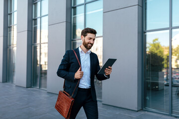 Glad handsome confident young caucasian guy with beard in suit chatting on tablet, goes to work