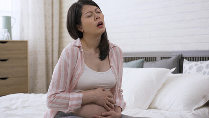 asian expectant mother suffering labor contraction is moaning and holding her tummy while her face...