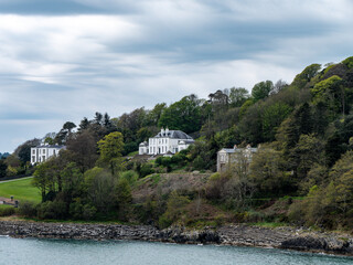 A hill on the shore of the bay. Several buildings and trees on the hill. Glandore is a village in the south of Ireland. Beautiful clouds, dramatic landscape. White building near trees and water - Powered by Adobe