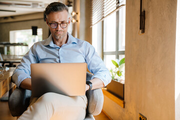 European grey man working with laptop while sitting in armchair at office