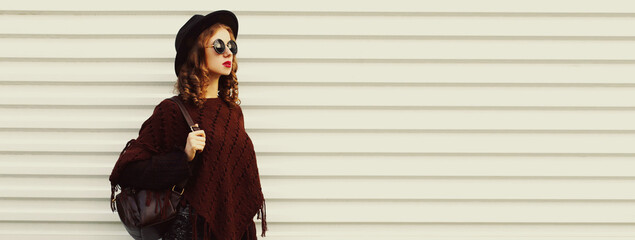 Portrait of stylish woman model posing wearing black round hat and knitted brown poncho on white...