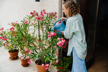 Young beautiful curly stylish woman in long shirt watering flowers