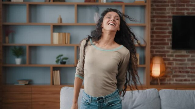 Happy active dancer girl housewife homeowner renter girl woman dancing in living room rejoice moving day relocation new home move head flying long curly hair looking at camera celebrate with dance