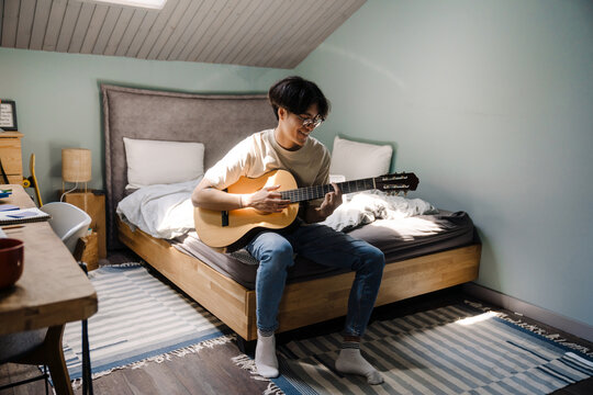 Young Asian Man In Headphones Playing Guitar While Sitting On Bed