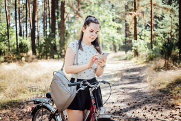 Fototapeta na wymiar Best cycling apps. Bike Tracker. Young woman with backpack riding bike and looking in cell phone on pine forest background. Girl with bike using a phone texting on smartphone app in forest walk