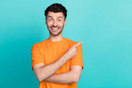 Photo of young handsome bearded bristle positive man wear orange t-shirt finger pointing empty space offer toothy beaming isolated on cyan color background