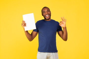 Cheerful Black Guy Holding Paper Gesturing Okay Over Yellow Background