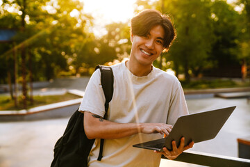 Young handsome smiling asian boy with backpack holding laptop