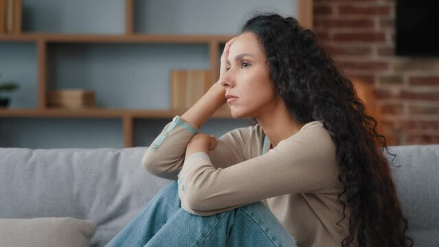 Pensive sad upset worried annoyed Hispanic Latino Caucasian woman 30s lady sitting on couch feeling sadness worry about problem stress trouble bad unwell headache unwanted pregnant bored mood indoors