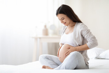 Smiling pregnant woman sitting on bed in her bedroom - obrazy, fototapety, plakaty