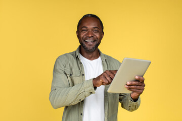 Happy surprised mature african american male in casual typing on tablet, isolated on yellow...