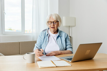 Fototapeta na wymiar A gray-haired, beautiful, stylish, serious lady works from home sitting at her desk with a laptop and notebooks for notes, in comfort