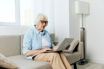 an elderly woman with white gray hair is sitting on a beige spacious sofa holding a laptop on her...