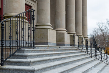 Marble steps lead to a large government building with five pillars or columns at the facade entrance, red door. The building has a large autumn tree in the background. The sky is cloudy and bright. 