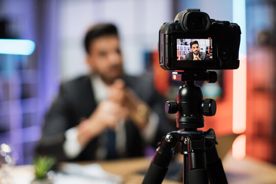 Focus on camera. Confident indian bearded businessman in suit sitting in front of camera in evening office during recording video for business vlog
