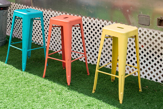 Three colorful iron metal stackable bar stools, blue, peach, and yellow color. The chairs are on green grass with white lattice in the background. The summer outdoor garden has shade and sunny spots.