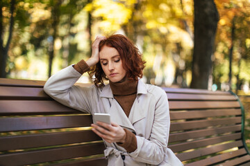 Unhappy caucasian millennial red-haired female in raincoat sits on bench, looks at smartphone in park