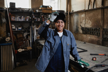Waist up portrait of multiethnic female welder smiling at camera in industrial factory workshop,...